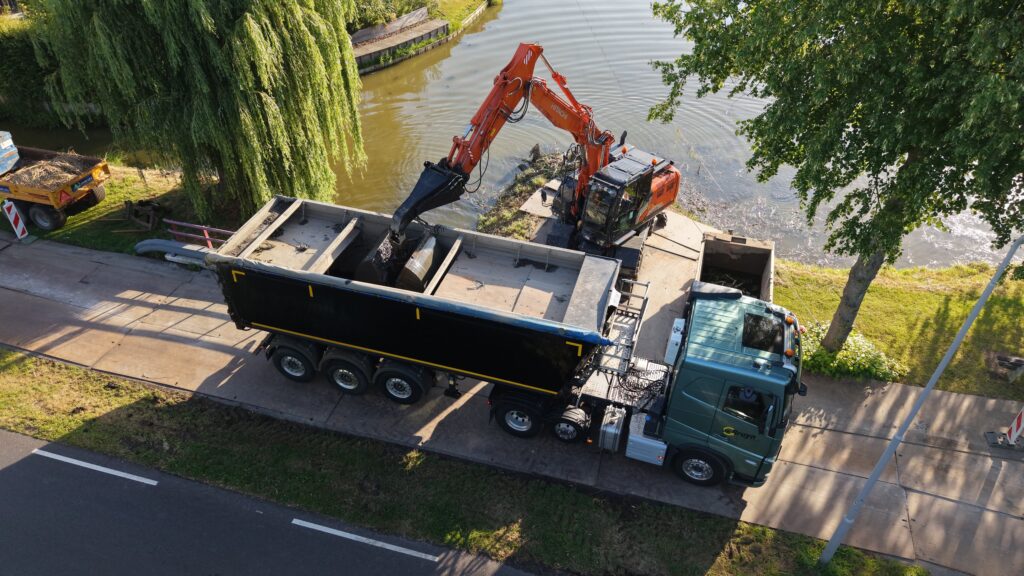 Vrachtwagen De Bruyn Transport voor baggertransport die met de kraan wordt geladen met slib uit de sloot bij een woonwijk.